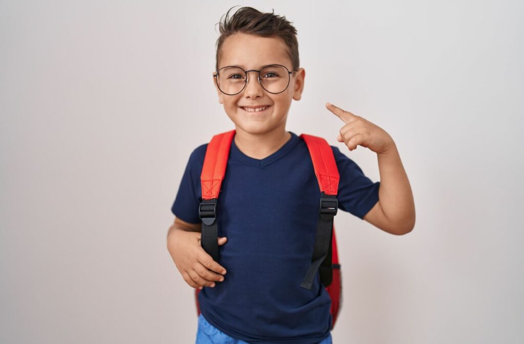 A young boy wearing a backpack happily points at his glasses