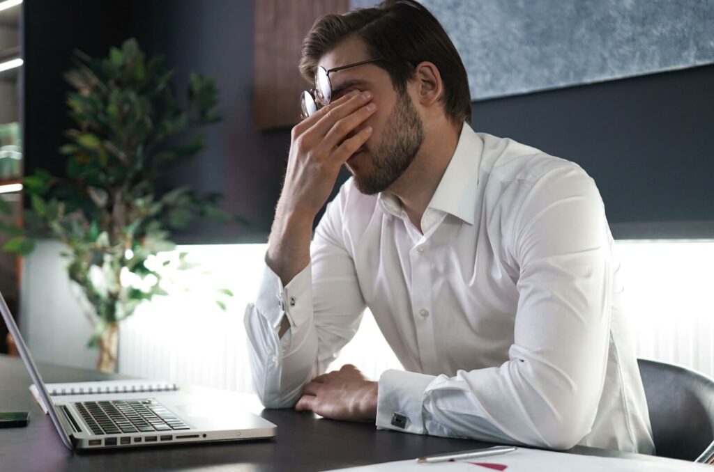 A person sits at their desk, rubbing their eyes with one hand while holding their glasses in the other.