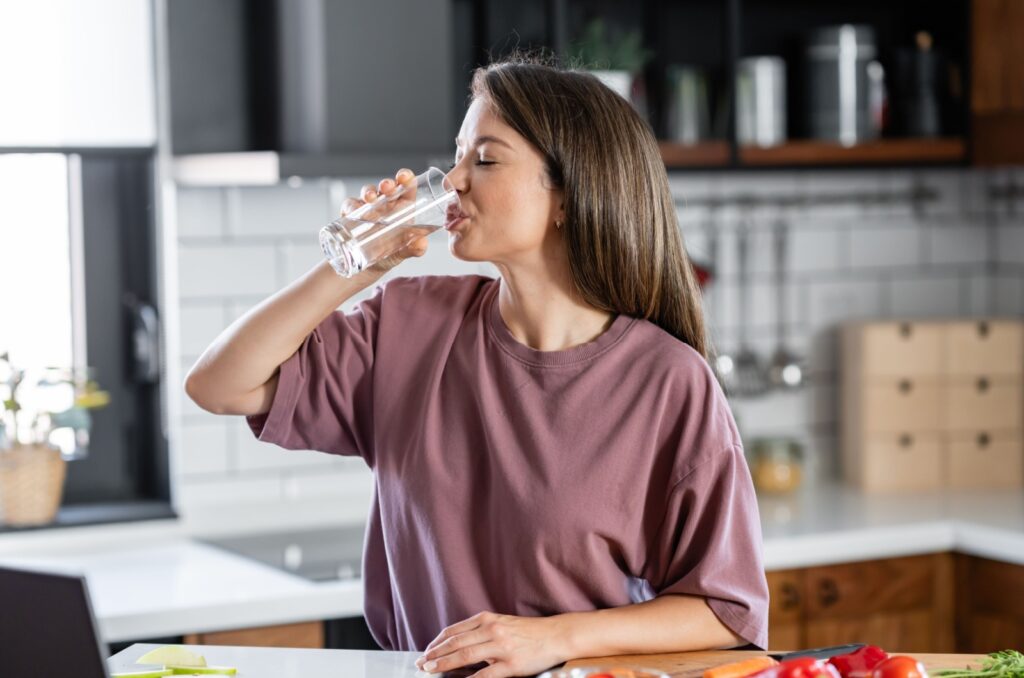 A person stands at their kitchen counter, drinking a glass of water.