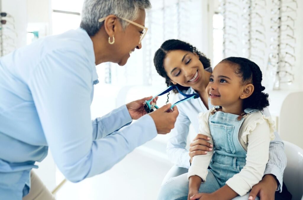 A young child sits with their parent in a brightly-lit optometrist’s office as their optometrist gives them a pair of glasses