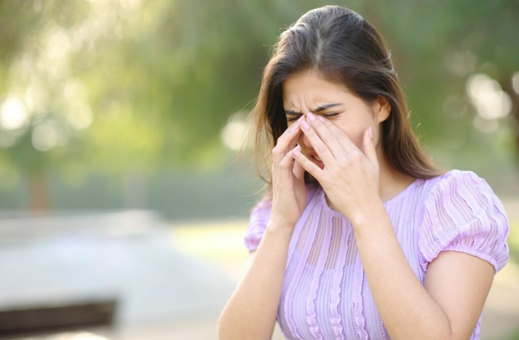 An adult in a purple shirt pinches the bridge of their nose to relieve allergy symptoms while outside in the daylight