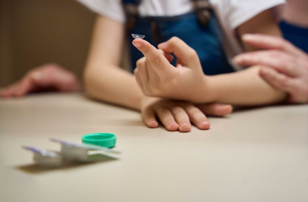 A child holds a contact lens on their fingertip while sitting at a table, with an adult and a lens case nearby.