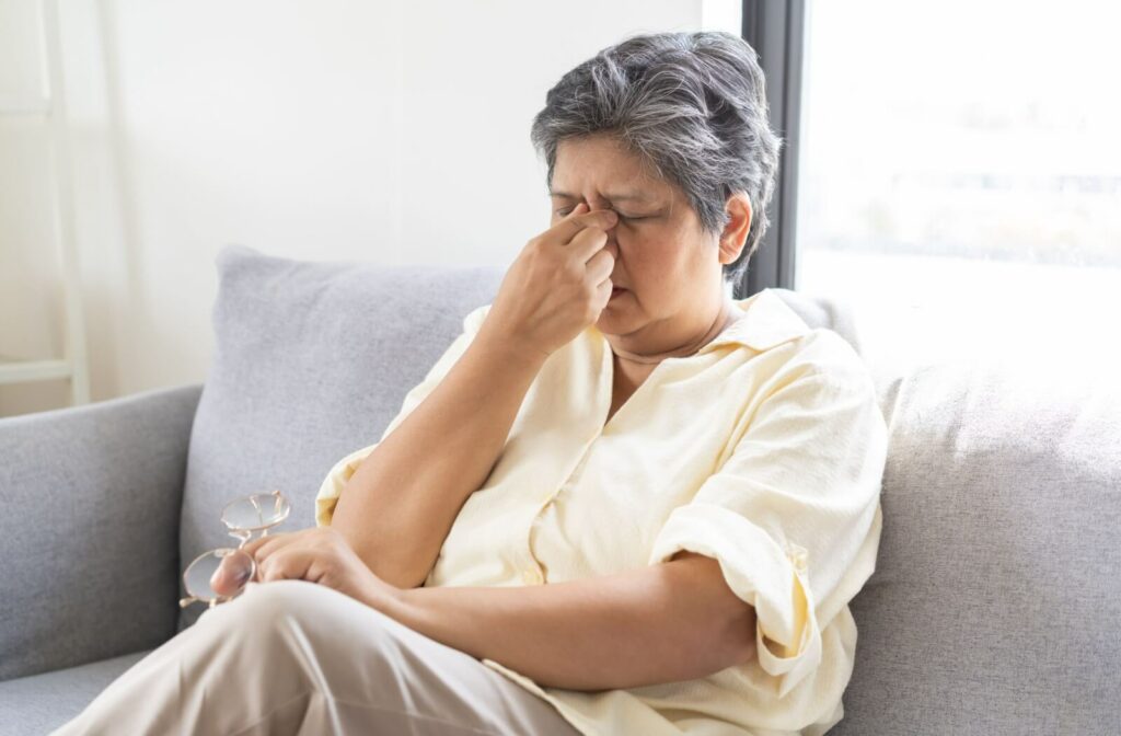 A person sits on a couch holding their glasses in one hand while rubbing the bridge of their nose, showing discomfort from dry eyes.
